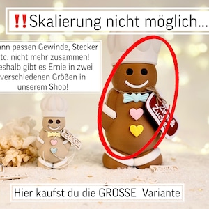 May include: A brown gingerbread man figurine wearing a chef's hat and bow tie, with pink and yellow heart accents. The figurine has a tag that says "Zucker". The text in the image says "Hier kaufst du die GROSSE Variante".