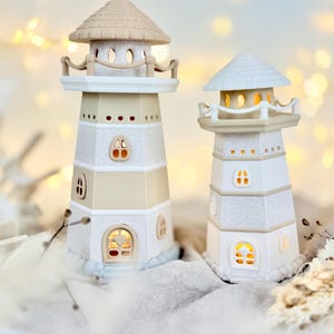 Op de afbeelding: Twee decoratieve vuurtorenbeeldjes. E&eacute;n is beige en wit, de andere is wit. Beide hebben kleine, verlichte ramen. De vuurtorens hebben een getextureerde, glinsterende afwerking en zijn bekroond met een klein dak.
