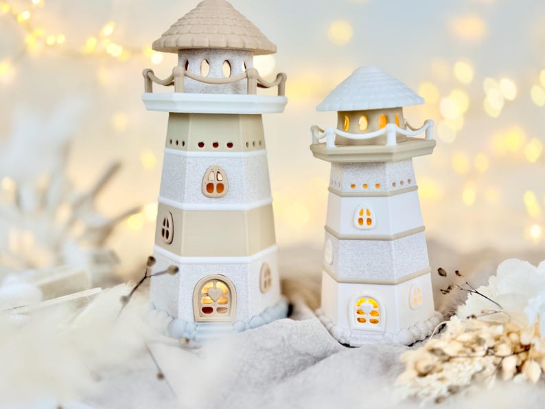 Op de afbeelding: Twee decoratieve vuurtorenbeeldjes. E&eacute;n is beige en wit, de andere is wit. Beide hebben kleine, verlichte ramen. De vuurtorens hebben een getextureerde, glinsterende afwerking en zijn bekroond met een klein dak.