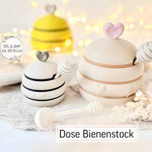 May include: Cream ceramic honey pots with heart-shaped lids and striped details. One pot is yellow with black stripes, resembling a bee. A matching spoon rests beside the pots. The text "Dose Bienenstock" is visible.