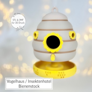 Puede incluir: Un comedero para pájaros gris claro y amarillo con forma de colmena. El comedero tiene una abertura redonda con un borde en forma de flor amarilla. La base es un plato amarillo con pequeños agujeros. El texto "Vogelhaus / Insektenhotel Bienenstock" está en la parte inferior.