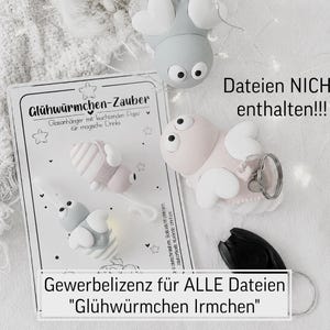 Könnte beinhalten: Eine Sammlung skurriler, hellfarbiger Dekorationsartikel, darunter drei kleine, stilisierte, insektenförmige Ornamente mit Flügeln und ein Schlüsselanhänger. Die Artikel sind auf einer weißen Oberfläche angeordnet. Der Text enthält die Worte "Glühwürmchen-Zauber".
