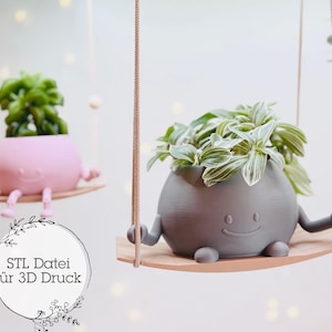 Puede incluir: Dos macetas colgantes impresas en 3D con caras sonrientes. La maceta de la izquierda es rosa y contiene una pequeña planta verde. La maceta de la derecha es gris y contiene una planta verde y blanca. El texto "STL Datei für 3D Druck" es visible en la imagen.