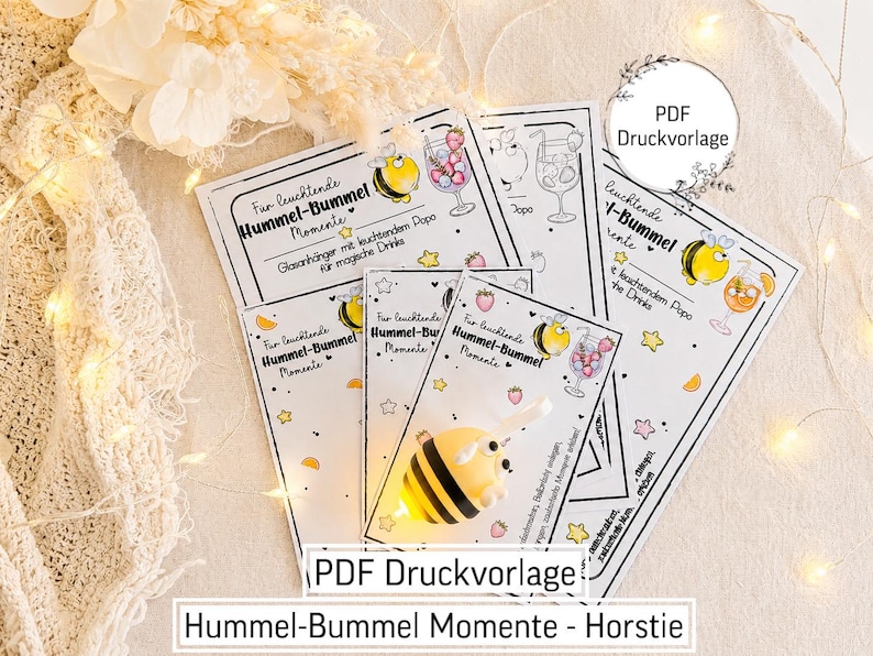 May include: A set of printed cards featuring illustrations of a bumblebee and a cocktail glass with the text "F&uuml;r leuchtende Hummel-Bummel Momente". A small, yellow and black striped bumblebee figurine with a glowing light is placed on top.