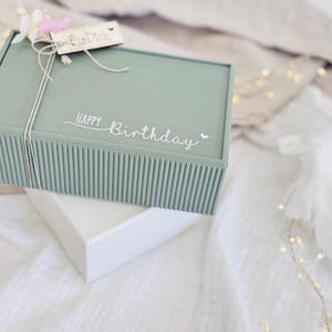 May include: A sage green gift box with a ribbed design, tied with twine and a small wooden tag. The words "Happy Birthday" are written on the box. The box is sitting on top of a white box.