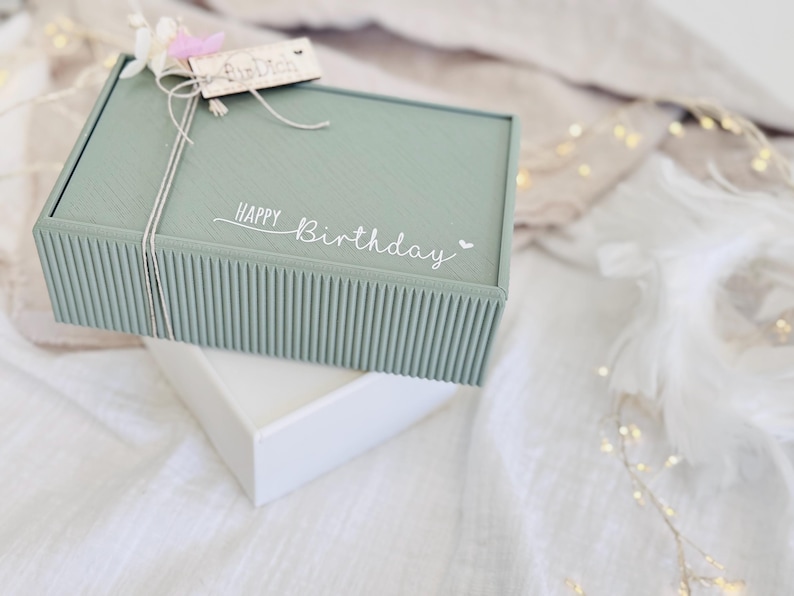 May include: A sage green gift box with a ribbed design, tied with twine and a small wooden tag. The words "Happy Birthday" are written on the box. The box is sitting on top of a white box.