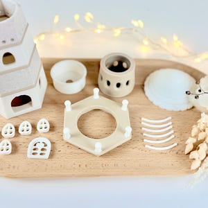 Op de afbeelding: Een verzameling beige en witte keramische stukken, waaronder een toren met meerdere niveaus, kleine ramen, een cirkelvormige container en een zeshoekig frame. Een witte zeeschelp en gedroogde bloemen zijn ook aanwezig, gerangschikt op een houten dienblad.