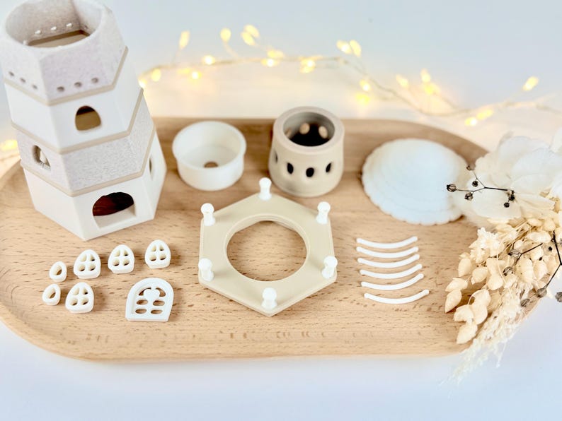 Op de afbeelding: Een verzameling beige en witte keramische stukken, waaronder een toren met meerdere niveaus, kleine ramen, een cirkelvormige container en een zeshoekig frame. Een witte zeeschelp en gedroogde bloemen zijn ook aanwezig, gerangschikt op een houten dienblad.