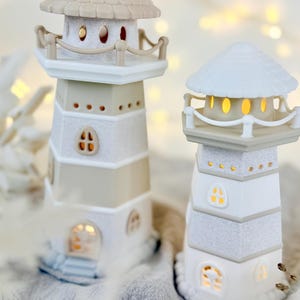 Op de afbeelding: Twee decoratieve vuurtorenbeeldjes in wit en beige. Elke vuurtoren heeft een gedetailleerd ontwerp met ramen en een verlicht interieur. De vuurtorens hebben een textuurafwerking en een nautisch thema.