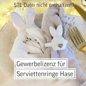 Op de afbeelding: Twee stoffen servetten, één wit en één beige, zijn versierd met witte konijnen servetringen. Elke konijnenring heeft een hartdetail. De servetten zijn versierd met takjes gedroogde bloemen. De tekst "Gewerbelizenz für Serviettenringe Hase" is zichtbaar.