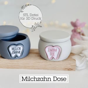 Pode incluir: Dois pequenos recipientes redondos com tampa, um cinzento e outro branco, assentam numa base de madeira. O recipiente cinzento tem uma tampa aberta e um emblema em forma de dente que diz "MILCH ZAHNE". O recipiente branco tem um emblema em forma de dente que diz "ZAHN FEE".