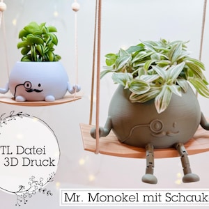 以下が含まれることがあります： 顔とモノクルが付いた3Dプリントされたプランターが2つ、スイングから吊り下げられています。プランターはライトブルーとグレーで、「STL Datei für 3D Druck」と「Mr. Monokel mit Schaukel」とラベル付けされています。