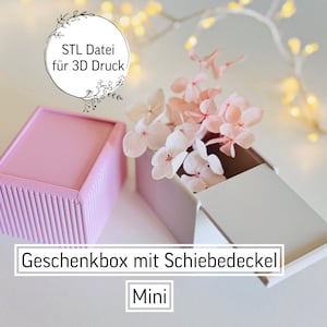 May include: A pink rectangular box with a lid, next to an open white box with pink flowers. The text "STL Datei für 3D Druck" is in a circle. The text "Geschenkbox mit Schiebedeckel" and "Mini" are also visible.