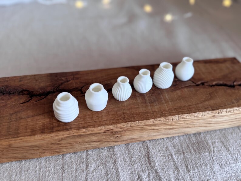 Pu&ograve; includere: Un set di sette piccoli vasi in ceramica bianca di forme e dimensioni diverse, disposti in fila su una superficie di legno.