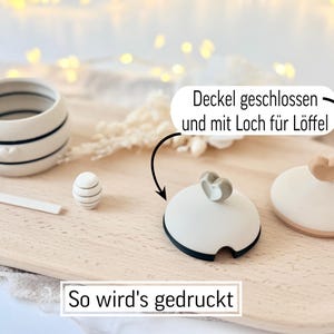 May include: A ceramic container with black stripes and a heart design, a small decorative egg, a spoon, and two lids with heart-shaped handles. The text "So wird's gedruckt" is at the bottom.