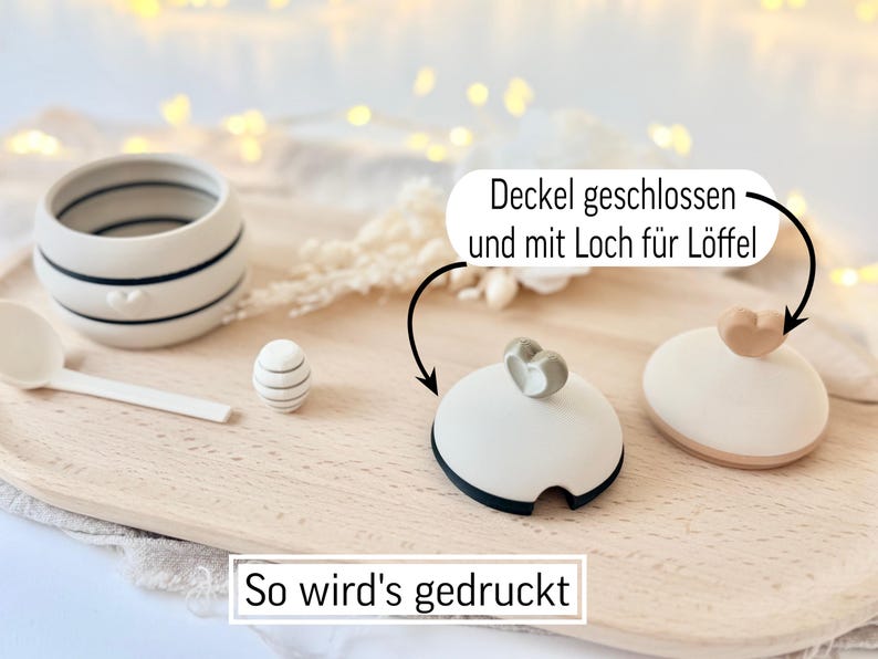 May include: A ceramic container with black stripes and a heart design, a small decorative egg, a spoon, and two lids with heart-shaped handles. The text "So wird's gedruckt" is at the bottom.
