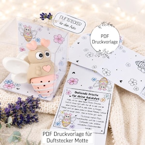 May include: A beige and pink moth-shaped air freshener with large eyes and heart details, packaged in a clear box. Included are printed paper cards with bee illustrations and German text, plus a sprig of dried lavender flowers.