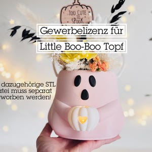 Könnte beinhalten: Ein rosa, geisterförmiger Pflanztopf mit schwarzen Augen und Mund, der einen weißen Kürbis mit einem leuchtenden Herzen hält. Der Topf ist mit Blumen gefüllt. Der Text auf dem Bild lautet "Too Cute to Spook" und "Gewerbelizenz für Little Boo-Boo Topf".