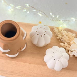 May include: A brown and white silicone tea infuser and two white flower-shaped tea infusers are arranged on a wooden tray. The infusers are designed for steeping loose leaf tea. Dried flowers and fairy lights are in the background.