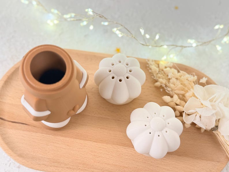 May include: A brown and white silicone tea infuser and two white flower-shaped tea infusers are arranged on a wooden tray. The infusers are designed for steeping loose leaf tea. Dried flowers and fairy lights are in the background.