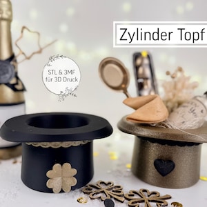 Puede incluir: Se muestran dos sombreros de copa en miniatura, uno negro con un trébol dorado y otro bronce con un corazón negro. Los sombreros están decorados con adornos dorados. La imagen también incluye una botella de champán y detalles decorativos. El texto "Zylinder Topf" es visible.