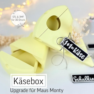 May include: A light yellow Käsebox, with the text "Käsebox" and "Upgrade für Maus Monty" in a white rectangle. A black tag with the text "1+1=Käse" is attached. The image also includes the text "STL & 3MF für 3D Druck" and "Schulkind".