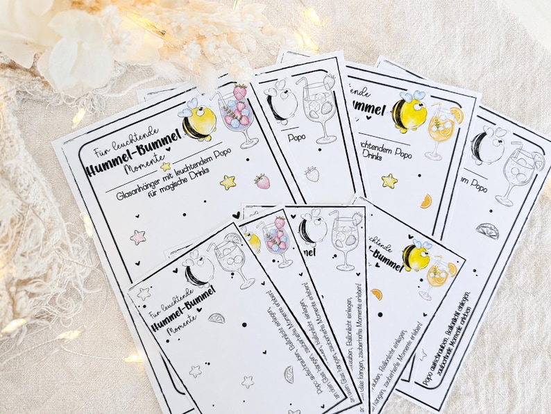 May include: A collection of rectangular white paper cards featuring black line art illustrations of bumblebees and cocktail glasses. The text reads "F&uuml;r leuchtende Hummel-Bummel Momente" and "Glasanh&auml;nger mit leuchtendem Popo f&uuml;r magische Drinks" with small yellow star and fruit accents.
