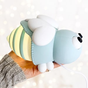 May include: A bee-shaped lamp with a light blue head, white wings, and a glowing yellow and light blue striped abdomen. The lamp features two black circular eyes and is connected to a thin white power cord extending from the bottom section.
