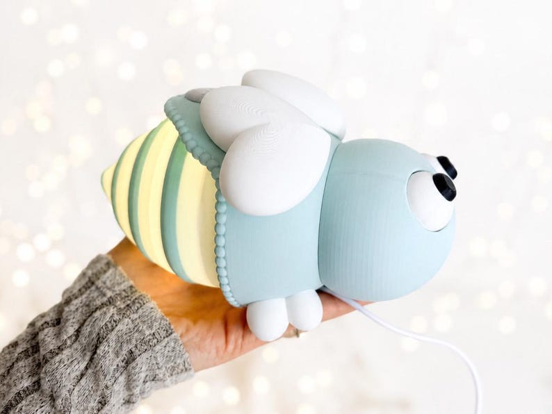 May include: A bee-shaped lamp with a light blue head, white wings, and a glowing yellow and light blue striped abdomen. The lamp features two black circular eyes and is connected to a thin white power cord extending from the bottom section.