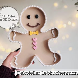 Puede incluir: Un plato decorativo con forma de hombre de jengibre con una cara sonriente, pajarita amarilla y botones en forma de corazón rosa. El plato está delineado con cuentas blancas. El texto en el plato dice "Dekoteller Lebkuchenmann".
