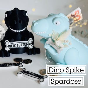 Może przedstawiać: Dwie skarbonki w kształcie dinozaurów, jedna czarna i jedna jasnoniebieska. Czarny dinozaur ma znak w kształcie kości z napisem "Bitte Füttern". Jasnoniebieski dinozaur ma kwiatową dekorację i pieniądze w szczelinie. Tekst "Dino Spike Spardose".
