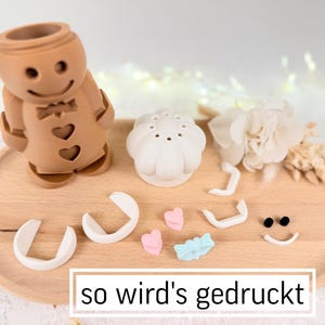 May include: A brown gingerbread man-shaped object with heart cutouts, a white flower, and other decorative elements on a wooden tray. The text "so wird's gedruckt" is at the bottom. Additional pieces include a white mushroom-shaped object, and small pink and blue shapes.