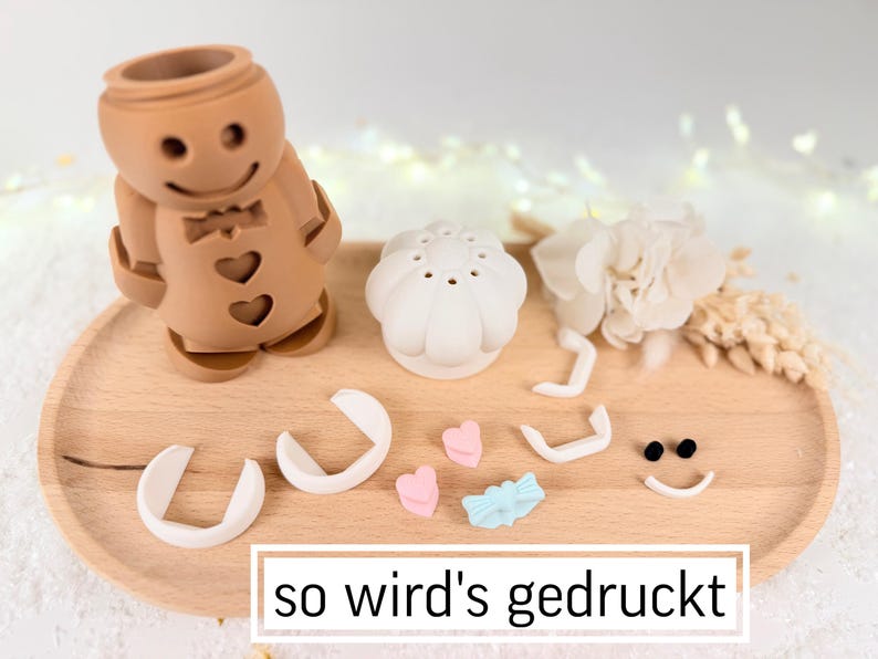 May include: A brown gingerbread man-shaped object with heart cutouts, a white flower, and other decorative elements on a wooden tray. The text "so wird's gedruckt" is at the bottom. Additional pieces include a white mushroom-shaped object, and small pink and blue shapes.