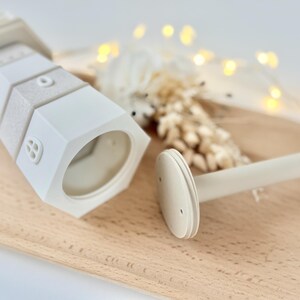 Op de afbeelding: Een wit en beige decoratief object in de vorm van een vuurtoren met een bijpassende staaf. De vuurtoren heeft een zeshoekige basis en getextureerde secties. De staaf heeft een cirkelvormig uiteinde. De items staan op een houten dienblad.