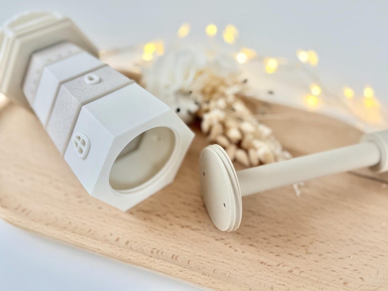 Op de afbeelding: Een wit en beige decoratief object in de vorm van een vuurtoren met een bijpassende staaf. De vuurtoren heeft een zeshoekige basis en getextureerde secties. De staaf heeft een cirkelvormig uiteinde. De items staan op een houten dienblad.