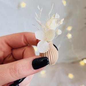 Pu&ograve; includere: Un piccolo vaso in ceramica beige con una texture a coste, riempito di fiori bianchi secchi e rami.