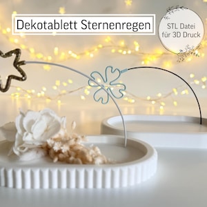 Può includere: Un vassoio decorativo bianco di forma ovale con bordo scanalato, contenente fiori secchi e un fiore bianco. Un design a filo a forma di stella e trifoglio è sospeso sopra il vassoio. Il testo "Dekotablett Sternenregen" è visibile.