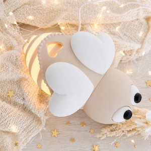 May include: A bee-shaped lamp with a beige body, white heart-shaped wings, and two black circular eyes. The lamp features a glowing light emanating from its striped abdomen and includes a white power cord extending from the top of the insect figure.