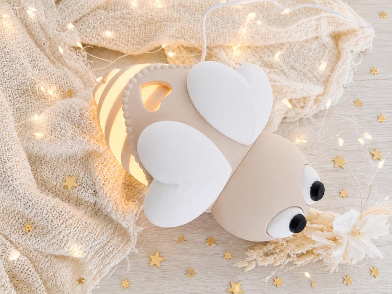 May include: A bee-shaped lamp with a beige body, white heart-shaped wings, and two black circular eyes. The lamp features a glowing light emanating from its striped abdomen and includes a white power cord extending from the top of the insect figure.