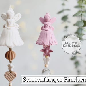 Puede incluir: Dos atrapasoles con temática de hadas. Uno es blanco, el otro rosa. Cada hada tiene alas y un vestido. El hada blanca tiene una cuenta marrón, una estrella y un corazón. El hada rosa tiene una flor, una mariposa y una cuenta de cristal. Texto: Sonnenfänger Finchen.