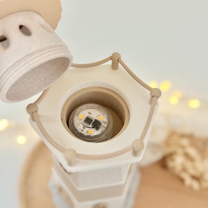 Op de afbeelding: Een beige en witte decoratieve lamp in de vorm van een vuurtoren met een afneembare bovenkant. De lamp heeft een gloeilamp aan de binnenkant en is ontworpen met een nautisch thema. De lamp is gemaakt van keramiek of een vergelijkbaar materiaal.