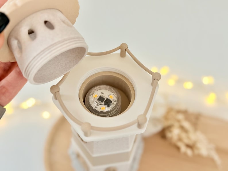 Op de afbeelding: Een beige en witte decoratieve lamp in de vorm van een vuurtoren met een afneembare bovenkant. De lamp heeft een gloeilamp aan de binnenkant en is ontworpen met een nautisch thema. De lamp is gemaakt van keramiek of een vergelijkbaar materiaal.