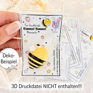 May include: A small yellow and black bumblebee-shaped charm attached to a white card with illustrations of strawberries, stars, and a cocktail glass. The card features the text "F&uuml;r leuchtende Hummel-Bummel Momente" and "Popo aufschrauben, Ball&ouml;nchen an dein Glas hangen, zauberhafte Momente".
