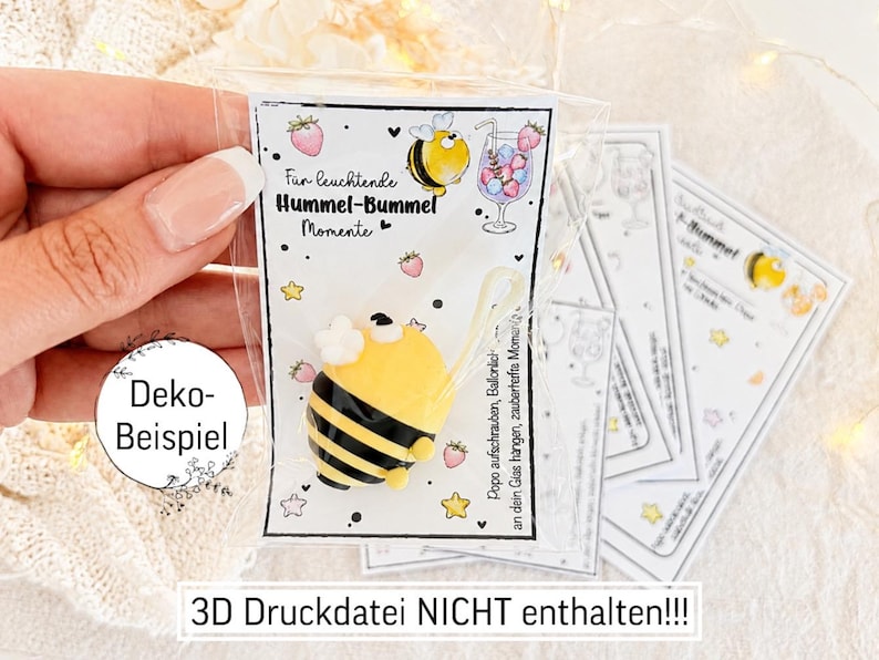 May include: A small yellow and black bumblebee-shaped charm attached to a white card with illustrations of strawberries, stars, and a cocktail glass. The card features the text "F&uuml;r leuchtende Hummel-Bummel Momente" and "Popo aufschrauben, Ball&ouml;nchen an dein Glas hangen, zauberhafte Momente".