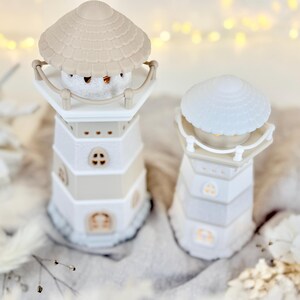 Op de afbeelding: Twee decoratieve kaarshouders in de vorm van een vuurtoren. De vuurtorens zijn wit en beige, met een schelpvormige bovenkant. E&eacute;n heeft een klein, rond, wit object binnenin. De andere heeft een lichtgekleurde, getextureerde bovenkant. De achtergrond is wazig met zachte verlichting.