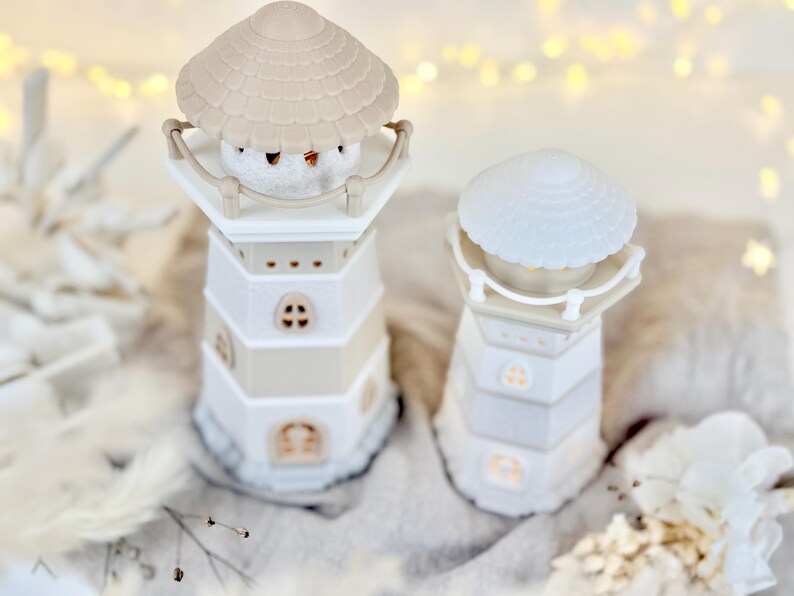 Op de afbeelding: Twee decoratieve kaarshouders in de vorm van een vuurtoren. De vuurtorens zijn wit en beige, met een schelpvormige bovenkant. E&eacute;n heeft een klein, rond, wit object binnenin. De andere heeft een lichtgekleurde, getextureerde bovenkant. De achtergrond is wazig met zachte verlichting.