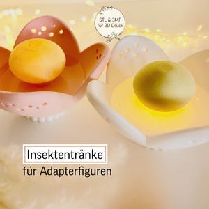 Puede incluir: Dos comederos para insectos con forma de flor, uno rosa y otro blanco, cada uno con una esfera amarilla. El comedero rosa tiene pequeños agujeros en los bordes. El texto de la imagen dice "Insektentränke für Adapterfiguren".