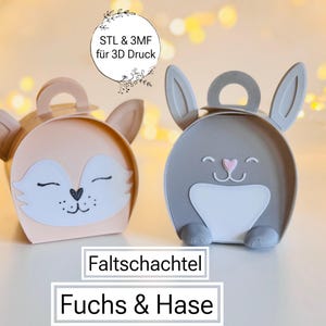 May include: Two decorative gift boxes shaped like animals. One is a peach-colored fox with a white face and black details. The other is a gray bunny with a white belly and pink details. The boxes have handles and are labeled "Fuchs & Hase".