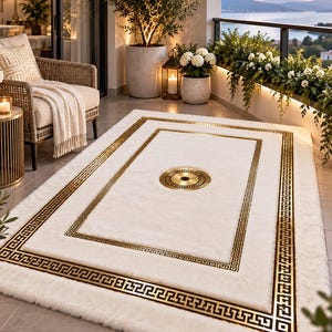 Puede incluir: Una alfombra rectangular con una superficie blanca y afelpada, que presenta un borde dorado con patrón de greca y un medallón circular dorado a juego en el centro, extendida sobre el suelo de baldosas de un balcón.