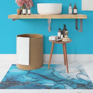 Puede incluir: Un ba&ntilde;o con una pared azul, un estante de madera con un lavabo blanco, una cesta de lavander&iacute;a de mimbre con una toalla blanca, una peque&ntilde;a mesa de madera con art&iacute;culos de tocador y una alfombra de ba&ntilde;o con estampado abstracto azul y blanco.